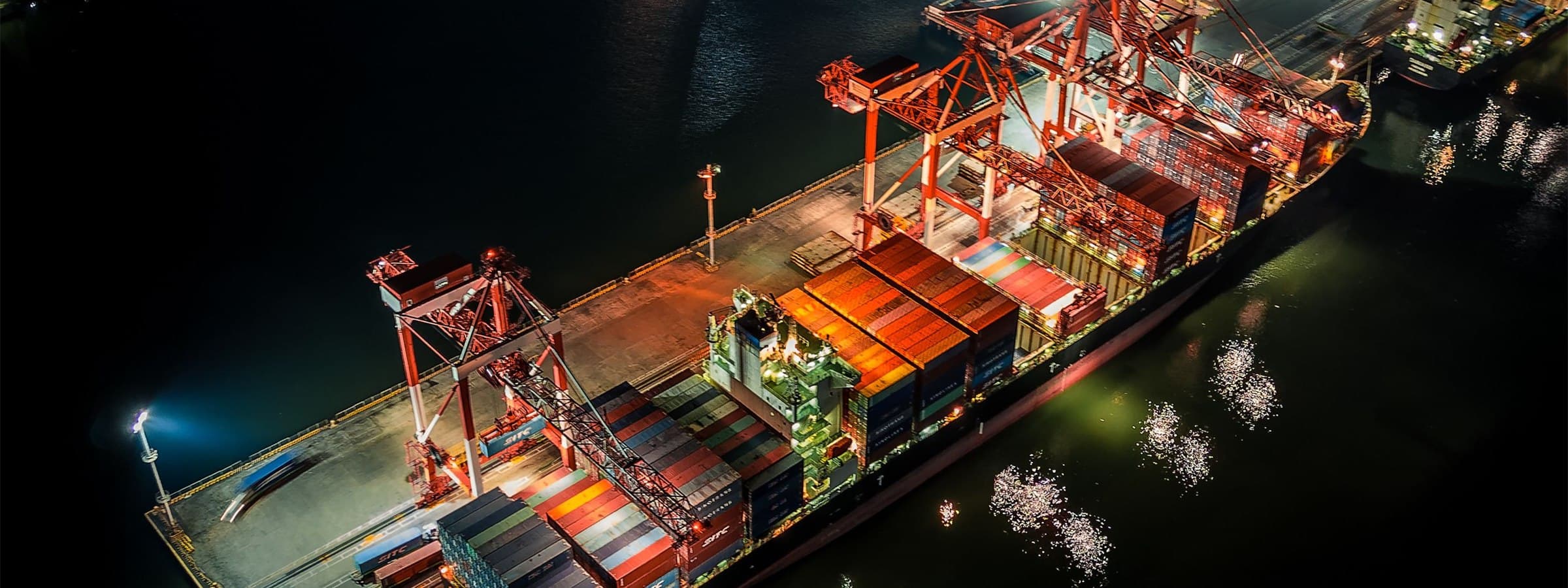 Aerial night view of a container vessel being worked alongside a lit terminal