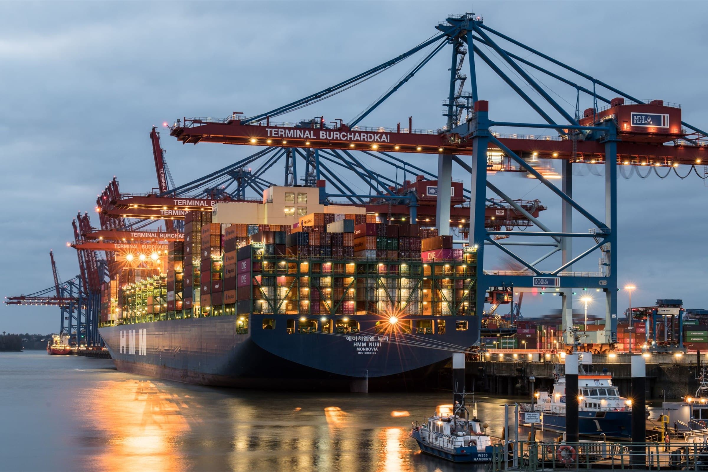A container terminal at dusk, cranes working a vessel alongside